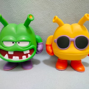 May include: Two cartoon character figurines. One is green with purple arms and legs and has a toothy grin. The other is yellow with orange arms and legs and wears purple sunglasses.