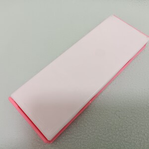 May include: A pink rectangular case with a white interior. The case is closed and lying flat on a light green surface.