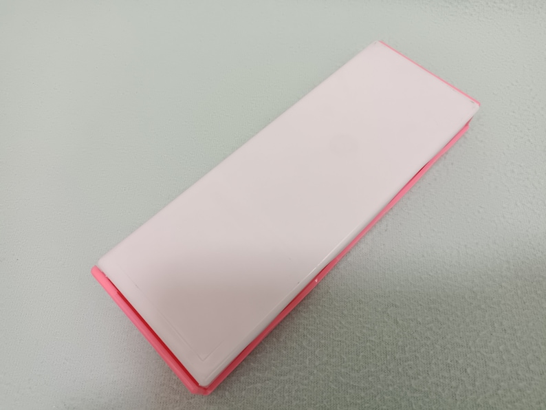 May include: A pink rectangular case with a white interior. The case is closed and lying flat on a light green surface.