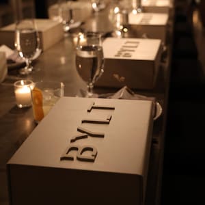 Peut inclure: Une série de boîtes rectangulaires beiges avec le mot BYLT en relief sur les couvercles, disposées en une longue rangée sur une table sombre. Chaque boîte est accompagnée d'un verre d'eau et d'une petite bougie allumée dans une pièce tamisée.