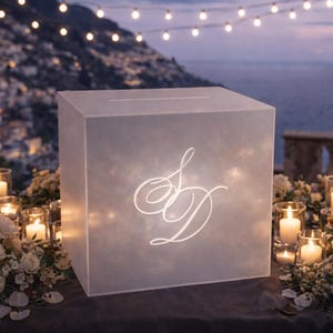May include: A softly illuminated white box, featuring the initials "S D" in a stylish script, with a slot for cards. The scene is set with candles, flowers, and fairy lights, creating a romantic atmosphere, perfect for a wedding or special event.