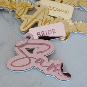 Puede incluir: Etiquetas de nombre de acr&iacute;lico color oro rosa con los nombres "Sam", "Bride", "Bridesmaid" y "Tiffany" escritos en letra cursiva. Las etiquetas est&aacute;n atadas a una cuerda.
