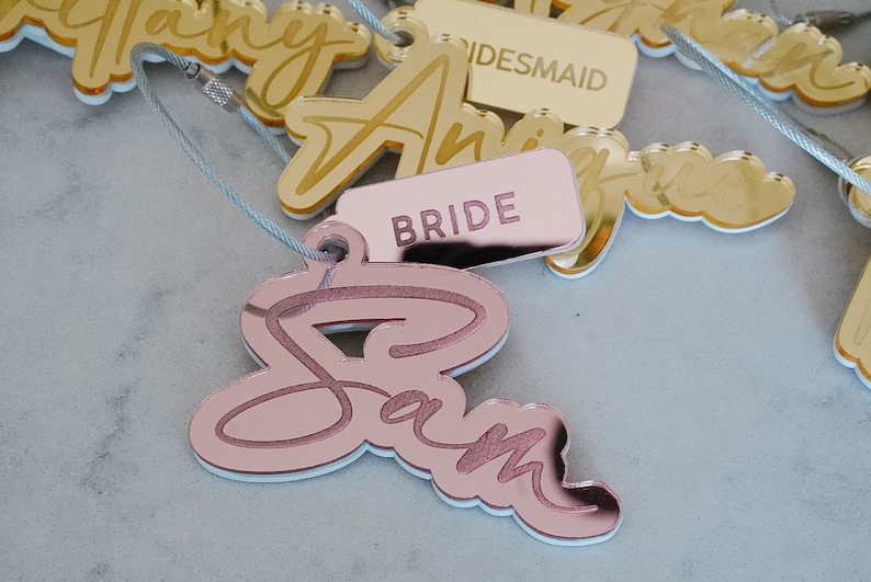Puede incluir: Etiquetas de nombre de acr&iacute;lico color oro rosa con los nombres "Sam", "Bride", "Bridesmaid" y "Tiffany" escritos en letra cursiva. Las etiquetas est&aacute;n atadas a una cuerda.