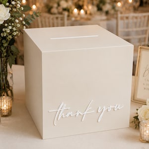 May include: A square, beige card box with a horizontal slot on the top surface. The front panel features the words "thank you" in a white, raised, cursive script font. The box has a smooth, matte finish and a clean, geometric design.