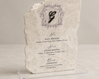Puede incluir: Una placa de piedra beige con bordes irregulares montada sobre una base de acrílico transparente. La placa presenta un marco decorativo con la letra G, seguido de recetas de cócteles para His Dirty Martini, Hers Lychee Martini y Ours L'Aperitivo.