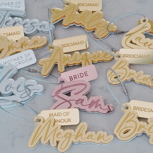 Puede incluir: Un conjunto de etiquetas de nombres de fiesta de boda de acr&iacute;lico dorado, plateado y rosa con una variedad de t&iacute;tulos que incluyen "Novia", "Dama de Honor", "Damas de Honor" y "Madre del Novio".