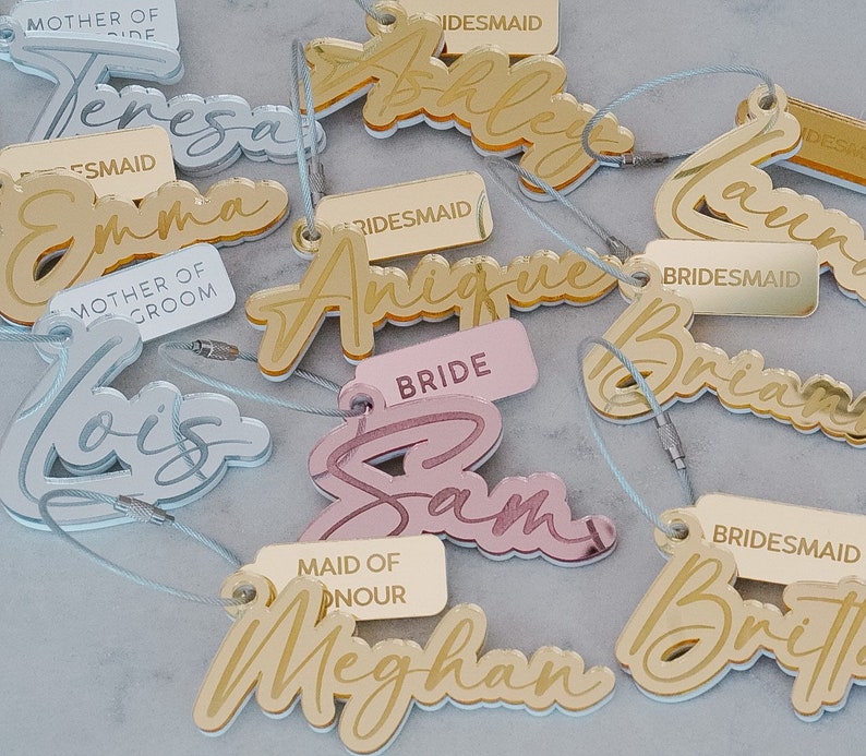 Puede incluir: Un conjunto de etiquetas de nombres de fiesta de boda de acr&iacute;lico dorado, plateado y rosa con una variedad de t&iacute;tulos que incluyen "Novia", "Dama de Honor", "Damas de Honor" y "Madre del Novio".