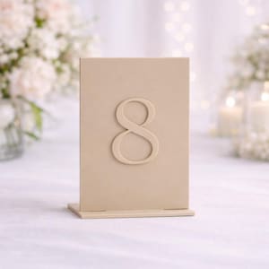 May include: A beige table number eight, ideal for weddings or events. The number is raised and centered on a rectangular card. The card sits on a small base. Soft focus background with flowers and candles.