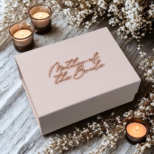 Può includere: Una scatola regalo rosa pallido con la scritta "Mother of the Bride" in corsivo. La scatola è circondata da fiori bianchi e candele accese in portacandele di vetro, creando un'atmosfera romantica.