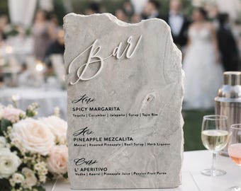 May include: A rectangular, rough-edged light gray stone slab sign featuring the white 3D text "Bar" at the top. Below, black printed text lists "Hers: Spicy Margarita", "His: Pineapple Mezcalita", and "Ours: L' Aperitivo" with ingredients.