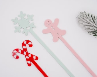 May include: Three festive metal drink stirrers with Christmas-themed designs: a red and white candy cane, a light blue snowflake, and a pink gingerbread person.