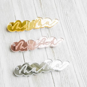 Puede incluir: Tres llaveros de acr&iacute;lico en forma de nube con la palabra "mama" escrita en blanco en cada uno. Los llaveros son dorados, de oro rosa y plateados.