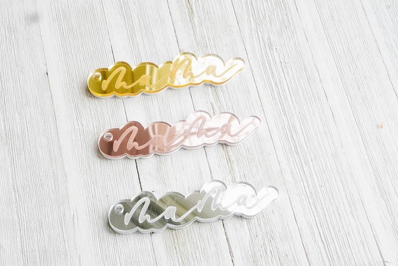 Puede incluir: Tres llaveros de acr&iacute;lico en forma de nube con la palabra "mama" escrita en blanco en cada uno. Los llaveros son dorados, de oro rosa y plateados.