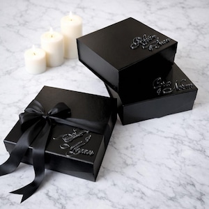 Könnte beinhalten: Schwarze Geschenkboxen mit geprägtem Schriftzug und Satinband. Eine Box ist geöffnet und gibt den Blick ins Innere frei. Im Hintergrund befinden sich drei weiße Kerzen. Auf den Boxen stehen die Worte "Refaire & Soin", "Cora & Norme" und "Faire & Soin".