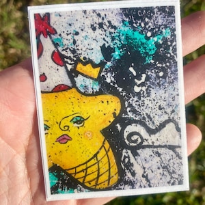 May include: A rectangular art print featuring a yellow face with a crown and a patterned design. The artwork has a white border and a background of black and white splatters with teal accents. The face has pink lips and black eyelashes.