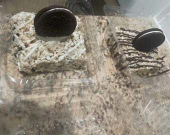 Pode incluir: Duas guloseimas crocantes de arroz com biscoitos Oreo em recipientes de plástico transparente. Uma guloseima é coberta com um biscoito Oreo e coberta com glacê branco. A outra é coberta com glacê de chocolate.