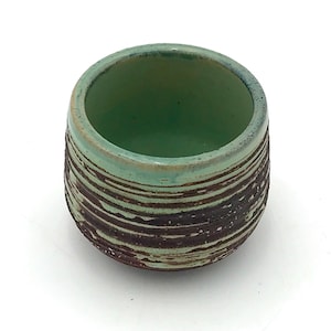 May include: A small, round ceramic cup featuring a smooth, light mint green interior and rim. The exterior displays a textured, dark brown finish with horizontal, uneven ridges that reveal hints of the light green glaze underneath.