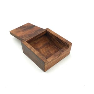 May include: A rectangular wooden box with a sliding lid that is partially open. The box features a rich, dark brown wood grain pattern throughout its surface, showing a smooth finish and visible natural wood textures on the interior and exterior.