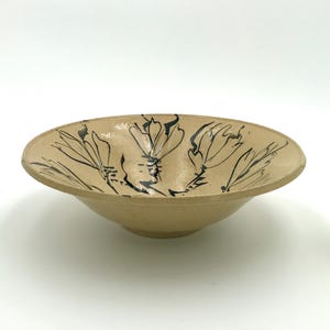May include: A beige ceramic bowl with a wide, shallow design. The bowl features a hand-painted floral pattern in black. The bowl is round with a slightly raised rim and sits on a white surface.