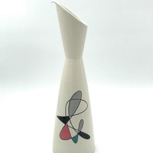 May include: A tall, off-white ceramic pitcher with a speckled texture and a slanted spout. The body features a graphic design consisting of thin black lines forming abstract loops, accented by solid shapes in muted pink, teal, and gray colors.