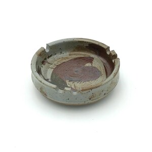 May include: A round ceramic ashtray with a speckled light gray glaze and a dark brown, circular brushstroke pattern in the center. The rim features three evenly spaced notches for resting items, and the overall finish has a smooth, glossy texture.