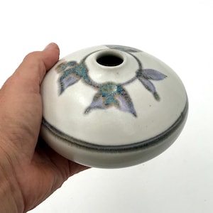 Może przedstawiać: Mały, okrągły wazon ceramiczny w kolorze złamanej bieli o gładkim, matowym wykończeniu. Górna część ozdobiona jest ręcznie malowanym motywem kwiatowym w odcieniach lawendy i błękitu, z ciemnoszarym poziomym pasem wokół dolnej części korpusu.