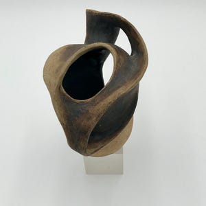 May include: A sculptural ceramic vessel featuring an abstract, twisted form with a dark, matte charcoal finish on the interior and a textured, earthy tan exterior. The object has an irregular, open-loop structure and rests on a clear base.