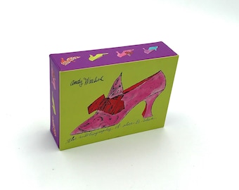 Peut inclure: Boîte rectangulaire avec un panneau avant vert citron représentant une illustration d'une chaussure à talon rose avec un nœud rouge. Le dessus et les côtés sont violets avec de petites illustrations. Le texte "Andy Warhol" et "the autobiography of alice b. shoe" sont visibles.