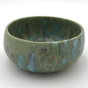 May include: A round ceramic bowl featuring a glossy glaze with a mottled pattern of sage green, muted teal, and soft brown tones. The surface displays a fluid, drip-like effect where the colors blend together across the exterior and interior.