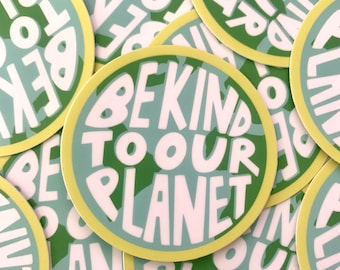 May include: A close-up of several round stickers with a green and white earth-toned background. The stickers have a yellow border and the text "Be Kind To Our Planet" in white.