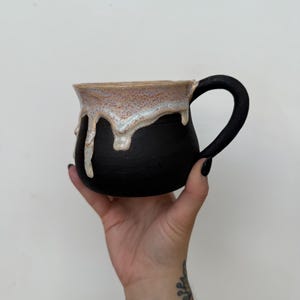 May include: A handmade ceramic mug with a matte black base and a contrasting glossy, light beige glaze dripping from the rim. The mug features a rounded body and a curved black handle, showcasing a distinct two-tone finish and texture.