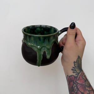 May include: A round ceramic mug with a matte black exterior and a glossy green glaze dripping from the rim down the sides. The mug features a dark handle and a smooth, rounded body, showcasing a contrast between the dark base and the vibrant green drip effect.