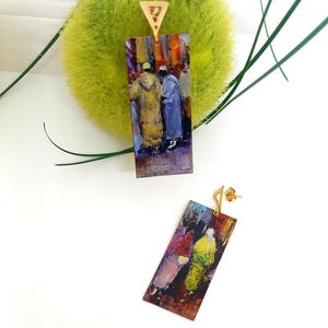 Può includere: Un paio di orecchini pendenti rettangolari caratterizzati da un colorato dipinto in stile impressionista di figure in abiti tradizionali. Ogni orecchino è sospeso a un piccolo gancio metallico triangolare color oro.