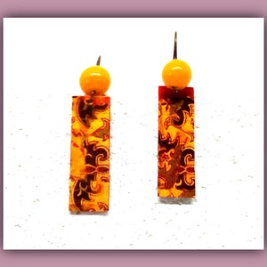 Peut inclure: Une paire de boucles d'oreilles pendantes avec une perle jaune en haut et un charme rectangulaire à motifs. Le charme est orange, rouge et noir avec un motif floral.