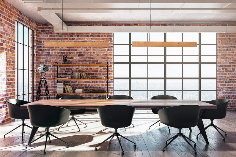 May include: A long wooden table with black chairs sits in a modern office space with exposed brick walls and large windows. The room has a minimalist design with a bookshelf and a tripod lamp.