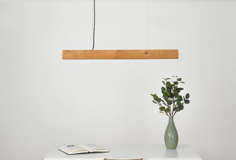 May include: A wooden pendant light fixture hangs from the ceiling. The light is made of a single, long, rectangular piece of wood. The light is hanging over a white table with a book and a vase of green leaves.