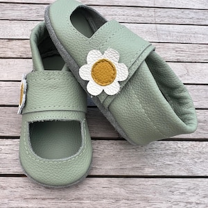 Könnte beinhalten: Ein Paar hellgrüne Babyschuhe im Mary-Jane-Stil. Jeder Schuh hat eine Applikation einer weißen und gelben Gänseblümchenblüte am Riemen. Die Sohlen sind grau, und die Schuhe sind aus einem weichen, strukturierten Material.