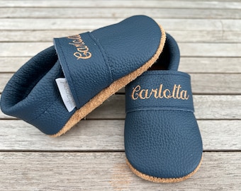 Puede incluir: Un par de zapatos de bebé de cuero azul marino con suela de gamuza. Los zapatos están personalizados con el nombre "Carlotta" bordado en hilo dorado.