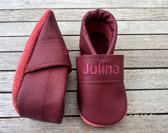 Op de afbeelding: Een paar baby-moccasins van bordeauxrood leer met roze stiksels. De mocassins hebben een zachte, suède voering en een flexibele zool. Eén moccasin heeft de naam "Julina" geborduurd op de band.