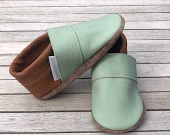 Op de afbeelding: Een paar lichtgroene leren slippers met bruine lederen afwerking en een suède zool. De slippers hebben een eenvoudig ontwerp en zijn perfect voor binnengebruik.
