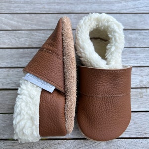Può includere: Un paio di pantofole in pelle marrone con fodera in pelliccia sintetica bianca. Le pantofole hanno un'etichetta "puschen power" sul lato.