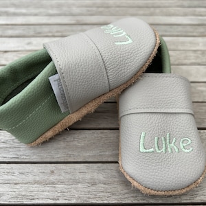 Pode incluir: Um par de sapatos de bebé em couro cinzento com o nome "Luke" bordado em verde claro. O interior é verde e as solas são de cor castanha natural. Sapatos para bebés.