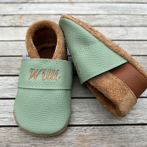 Op de afbeelding: Baby slofjes van mintgroen en bruin leer, met de naam "Willi" geborduurd in goudkleurig garen. De schoentjes hebben een zachte suède binnenkant en een leren bandje. Ontworpen voor baby's en peuters.