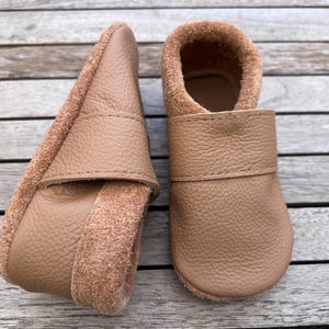 Op de afbeelding: Een paar bruinleren babyschoenen. De schoenen hebben een zachte, suède-achtige binnenkant en een gladde, getextureerde buitenkant. Het ontwerp omvat een band over de bovenkant en een afgeronde neus. De schoenen zijn ontworpen voor baby's en peuters.