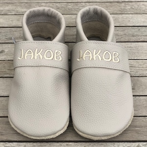 Könnte beinhalten: Ein Paar graue Leder-Babymoccasins mit dem Namen "JAKOB" auf jeder Schuh.