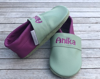 Op de afbeelding: Twee paar zachte leren baby-moccasins met paarse stiksels. Het ene paar is groen met de naam "Anika" geborduurd op de voorkant. Het andere paar is paars met de naam "Julie" geborduurd op de voorkant.
