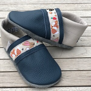 Op de afbeelding: Een paar marineblauwe en grijze babyschoenen. De schoenen hebben een band met een bosdierenpatroon met vossen, konijnen en paddenstoelen. De schoenen zijn gemaakt van leer en hebben een zachte zool.