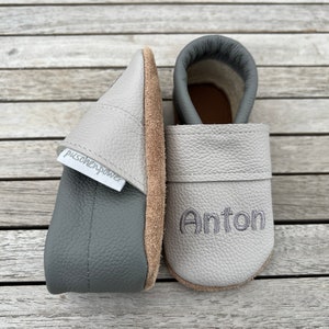 Op de afbeelding: Een paar grijze leren babyschoentjes met de naam Anton geborduurd op de ene schoen en de merknaam "puschenpower" op de andere.