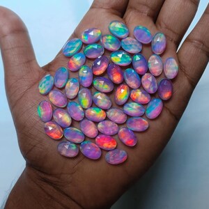 Op de afbeelding: Een verzameling van regenboogkleurige ovale edelstenen in tinten roze, paars en blauw. De edelstenen zijn op een hand van een persoon in een stapel gelegd.