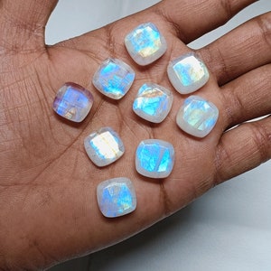 Peut inclure: Une collection de dix cabochons de pierre de lune blanche iris&eacute;e, chacun avec un motif unique de reflets bleus et dor&eacute;s. Les pierres sont pr&eacute;sent&eacute;es sur une main.